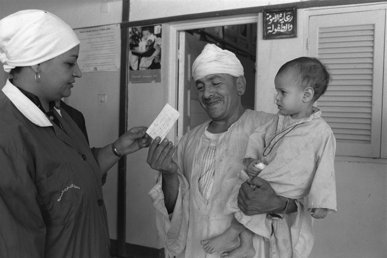 Carrying his toddler son, a man smiles as he hands a health card to a nurse at a UNICEF-assisted health centre.