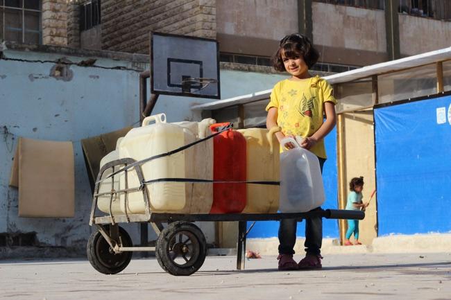 Huda stands next to a cart holding jerry cans filled with water.