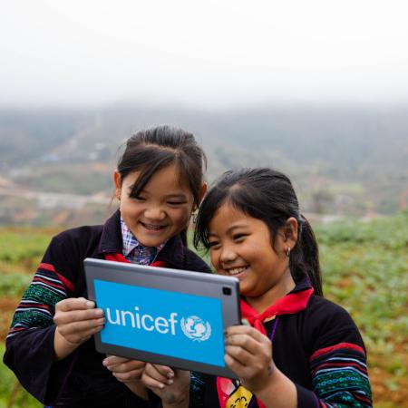 Two girls stand outdoors in a mountainous green field, looking at a tablet with a UNICEF logo on it. This photo was taken in Vietnam. 