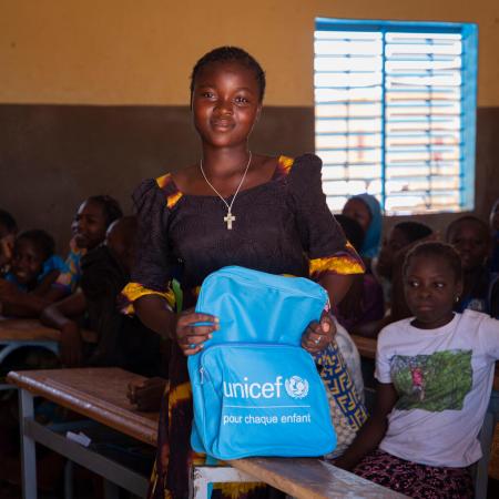 A girl hold a blue UNICEF backpack in front of a classroom full of students.