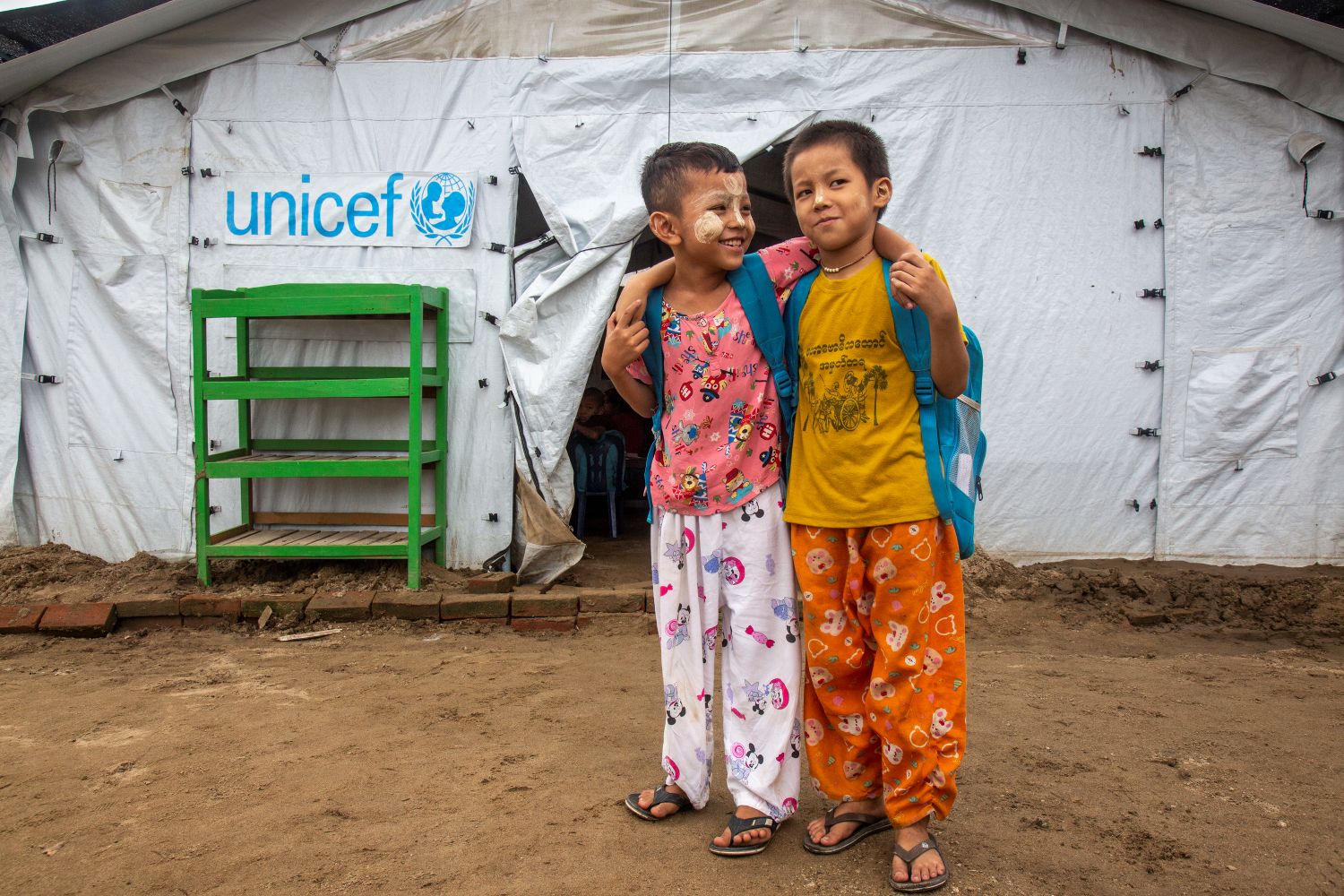 Two children stand outside a temporary UNICEF tent, smiling and with arms around each other, wearing backpacks.
