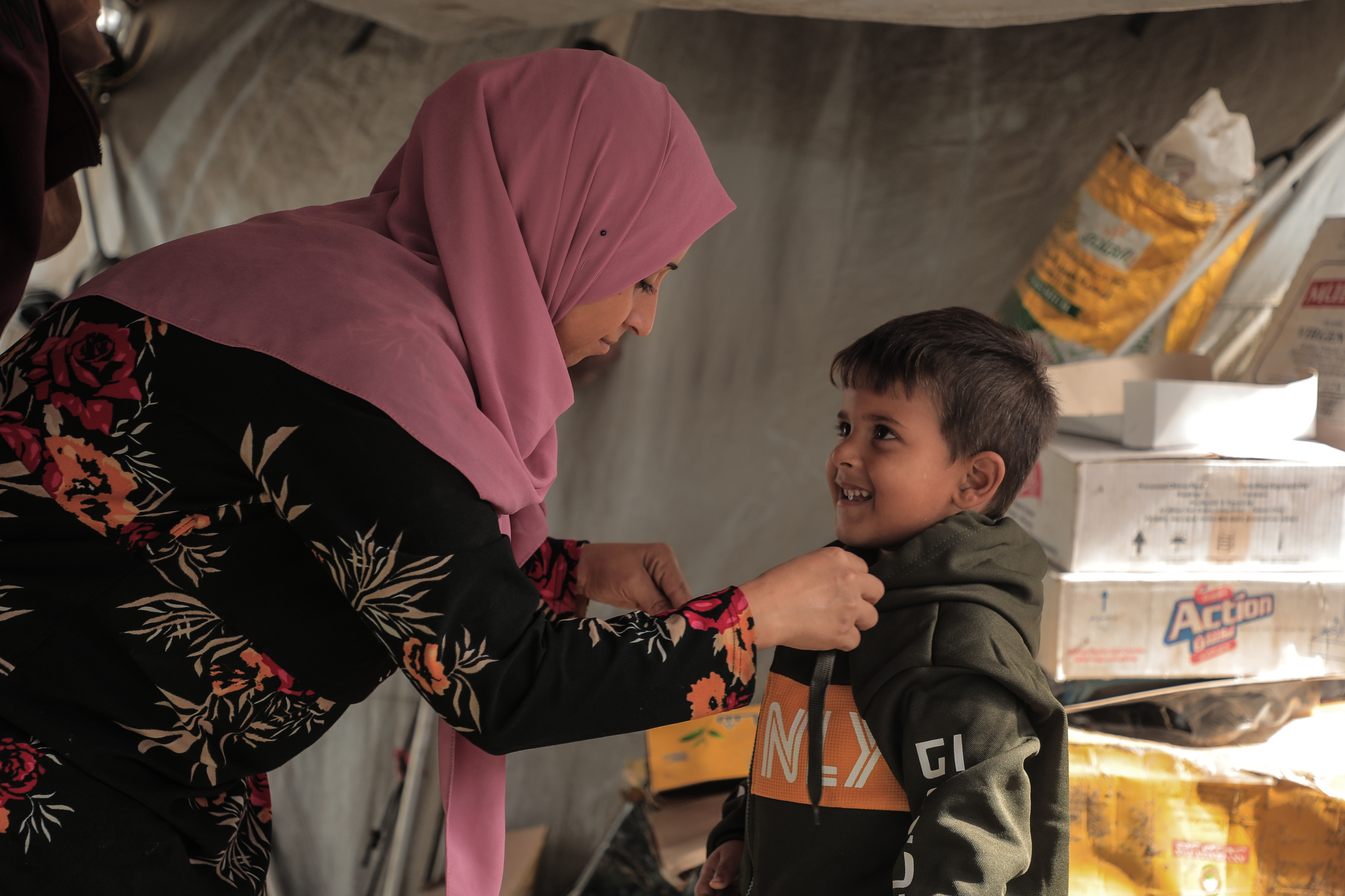 A smiling child looks up at his mother as she helps him put on a warm jacket.