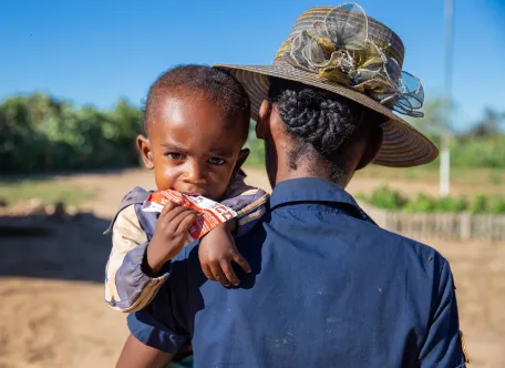 A boy being carried by his mother looks at the camera while eating a packet of therapeutic food called Plumpy’Nut.