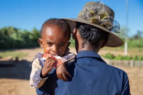 A boy being carried by his mother looks at the camera while eating a packet of therapeutic food called Plumpy’Nut.
