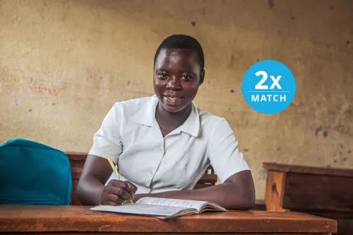Portrait of a student sitting at a school desk holding a pencil over an exercise book and looking at the camera with a neutral expression, with the overlaid copy 2X Match.