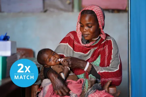An infant looks calmly at the camera while being held on an adult’s lap and eating from a packet of therapeutic food. Overlaid text says 2X Match.