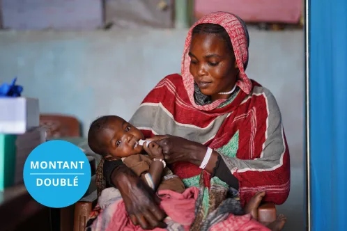 Un jeune enfant en train de manger le contenu d’un sachet d’aliment thérapeutique regarde calmement l’objectif, assis sur les genoux d’une adulte. Le texte superposé indique « Montant doublé ».