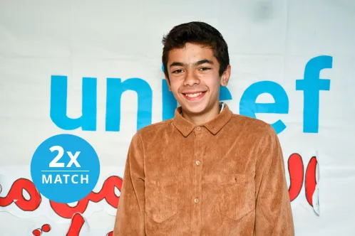 Portrait of a young child smiling at the camera in a sunny outdoor setting in front of a clay building.