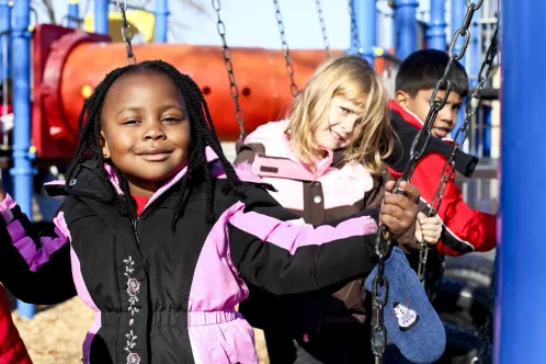 Three children from diverse backgrounds sit on swings in a park. 