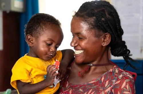 Une femme sourit au jeune enfant qu’elle tient dans ses bras. L’enfant tient un sachet d’aliment.