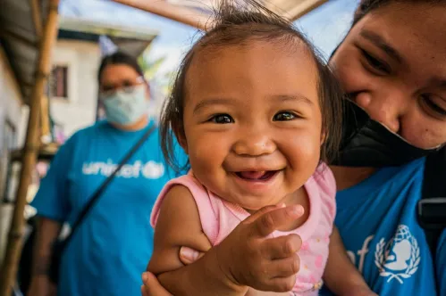 Close up of a smiling toddler girl with a UNICEF worker behind her. 