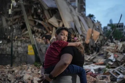 A child being carried by an adult in front of a collapsed building.