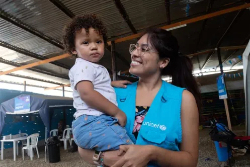 Une femme portant une veste bleue de l’UNICEF porte un enfant dans ses bras.