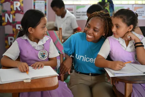 In a classroom, a woman wearing a blue UNICEF shirt crouch and smiles beside two girls sitting at their desk.