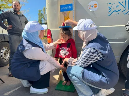 A child standing and having their height measured by two health workers at a height chart that has the UNICEF logo on it.