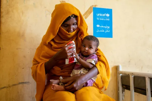 Against a yellow wall with a UNICEF sign on it, a woman looks down at a baby sitting on her lap who is looking at the camera and reaching for a food packet in the woman’s hands.