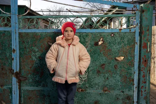 En Ukraine, une jeune fille portant un bonnet rouge se tient devant un portail métallique percé d’éclats d’obus.