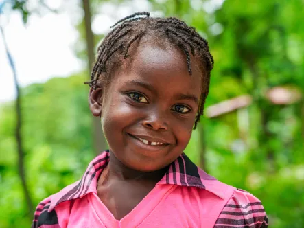 Close-up portrait of a child wearing short dreadlocks and a bright pink sports jersey and smiling at the camera against a backdrop of green foliage.