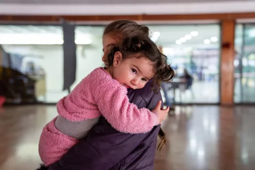 A mother holds a toddler wearing pink clothes in her arms in a brightly lit room.