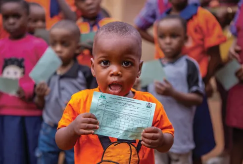Portrait of a child wearing an orange t-shirt, standing in front of a group of children and looking directly at the camera while holding a piece of paper below their chin.