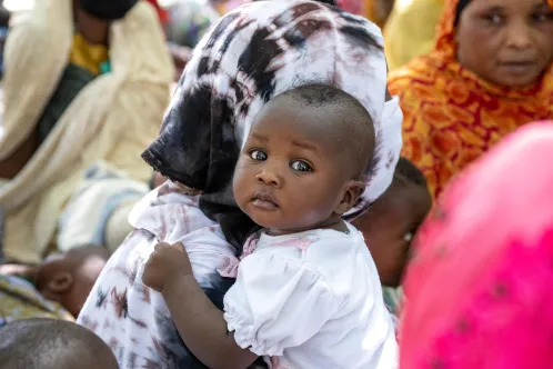 A baby being held by a woman wearing a white headscarf looks at the camera.
