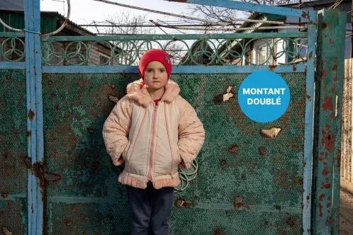 En Ukraine, une jeune fille portant un bonnet rouge se tient devant un portail métallique percé d’éclats d’obus.