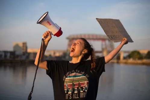 A girl with a megaphone in one hand and a poster in her other hand, speaks into the megaphone.