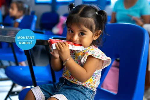 Portrait en gros plan d’une jeune enfant assise sur une chaise et mangeant le contenu d’un sachet d’aliment thérapeutique tout en regardant l’objectif.