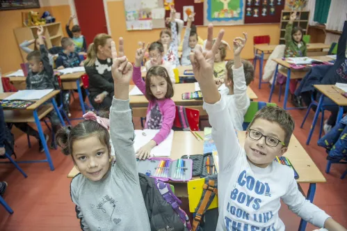 A classroom full of elementary school children, raise their hands to signal a V sign to encourage voting.