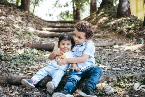A young boy sits in the woods, embracing his little sister from behind. 