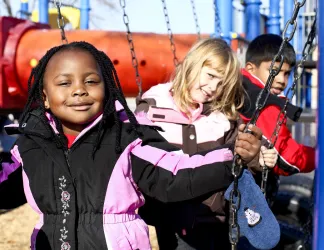 Trois enfants d’horizons différents sont assis sur des balançoires dans un parc.