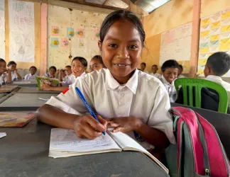 A smiling girl in a white school uniform writes in her notebook at a desk, with classmates seated behind her in a classroom decorated with students’ work.