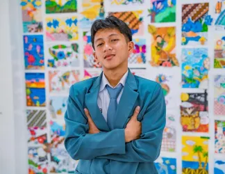 A teenage boy in a blue school blazer and tie stands with his arms crossed in front of a wall covered in colourful student artwork.