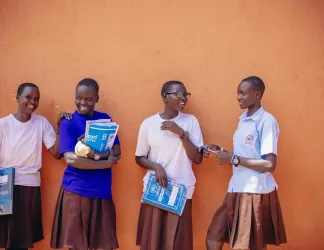 Four smiling schoolgirls in brown skirts lean against an orange wall, holding notebooks and chatting with one another.