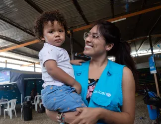 UNICEF worker in a blue vest smiling at a toddler she's holding.