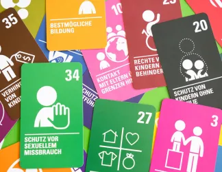 Colourful playing cards scattered on a green surface, each showing a simple white icon, a number and a German title for an article of the UN Convention on the Rights of the Child, including education, protection from sexual abuse, food, clothing and a safe home, and the best interests of the child.
