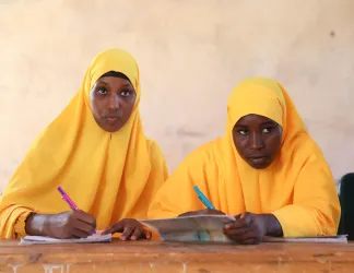 Two girls in yellow hijabs sit at a worn wooden desk in a classroom, pens in hand, writing in notebooks and looking toward the front of the room.