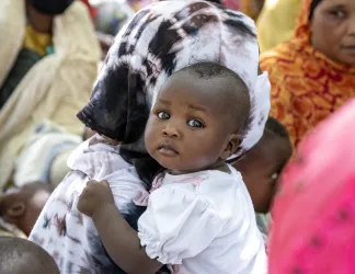 Baby looking at the camera while held by a woman in a patterned headscarf, in a crowd.