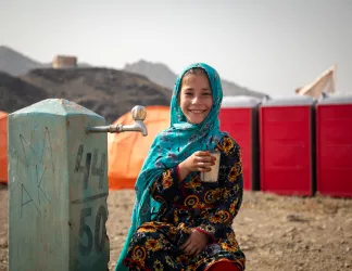 Girl in a headscarf smiling and holding a metal cup next to a water tap.
