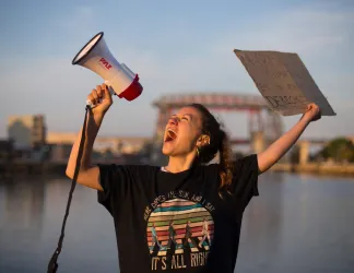 A girl with a megaphone in one hand and a poster in her other hand, speaks into the megaphone.