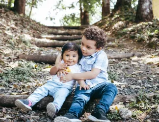 A young boy sits in the woods, embracing his little sister from behind. 