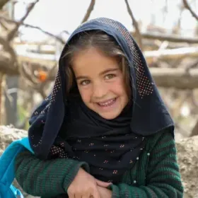 Smiling young girl in a black, sparkly headscarf and green sweater poses outdoors among bare tree branches, conveying warmth and joy.