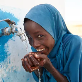 Girl in a headscarf drinking water cupped in her hands from a tap.