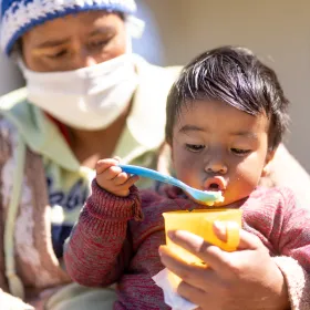 Toddler eating with a spoon, watched by a caregiver wearing a face mask.