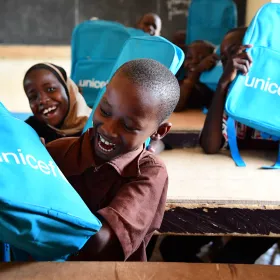 Children in a classroom laughing while holding turquoise UNICEF backpacks.