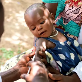 Baby receiving a vaccination in the arm outdoors, held by a caregiver.