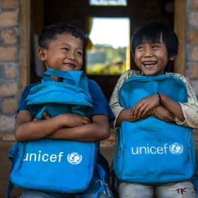 Two children laughing while holding UNICEF backpacks in front of a brick building.