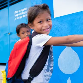 Girl with a backpack washing her hands at an outdoor tap, smiling at the camera.