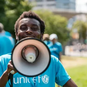 Smiling UNICEF volunteer in a blue shirt speaks through a megaphone outdoors, with a community gathering, lawn, trees, and city buildings behind.