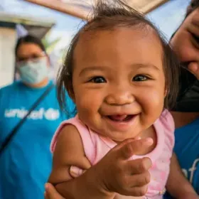 Smiling baby girl in a pink shirt is held by an adult, with a masked caregiver in blue behind her; warm, joyful family moment.